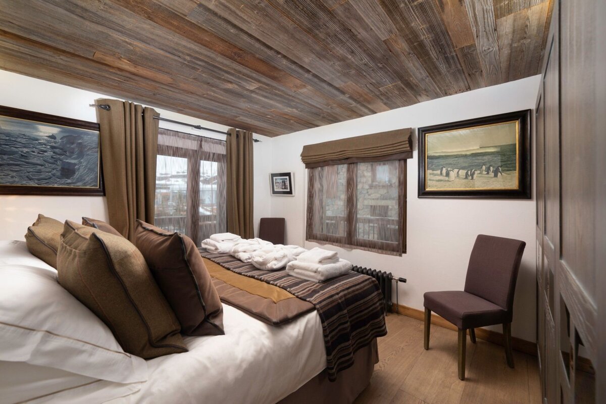 Cozy bedroom with a rustic wood-paneled ceiling, large bed with brown accents, two framed paintings, and windows with sheer brown curtains.
