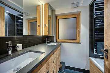 A modern bathroom features a wooden double vanity with two sinks, dark tiled backsplash, and a frosted window. Illuminated mirrors add a warm touch.