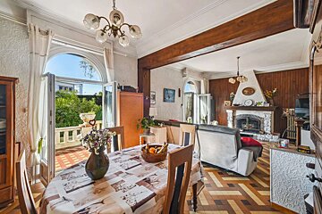 An inviting open-plan living/dining area with a rustic fireplace, wood paneling, and chevron tiled floor. French doors lead to a sunlit patio and garden.