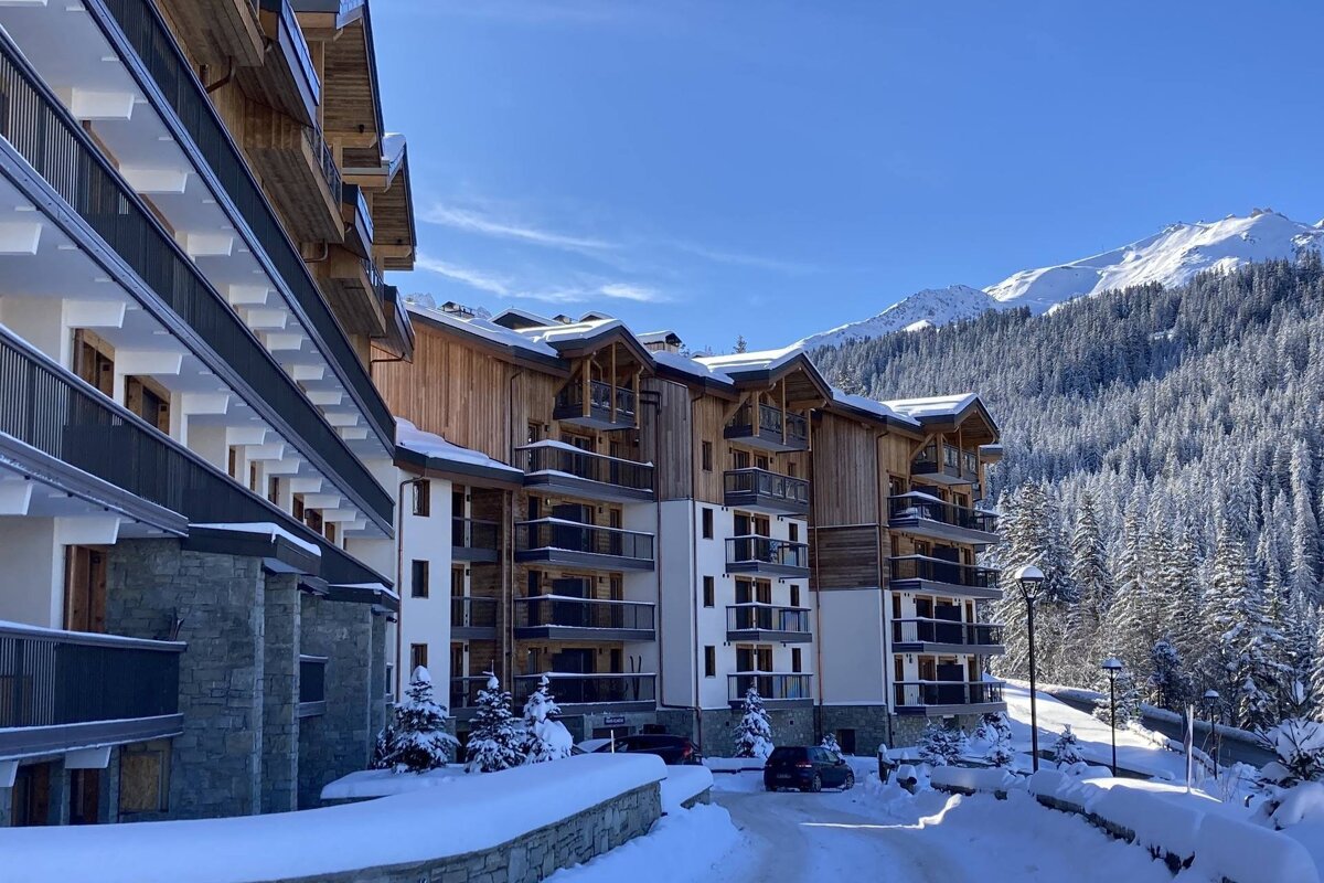 Snow-covered wooden alpine resort buildings set against a backdrop of snow-capped mountains and pine forests under a clear blue sky.