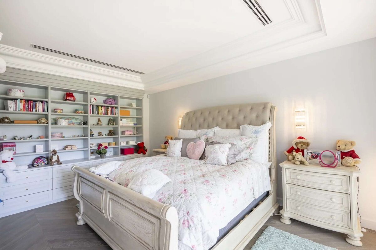 A spacious, elegant bedroom with a white upholstered bed, floral quilt, extensive built-in bookshelves, and playful teddy bears.