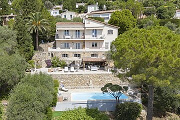 A scenic multi-story hillside villa with terraced stone walls, a swimming pool, and patio seating, surrounded by lush green trees.