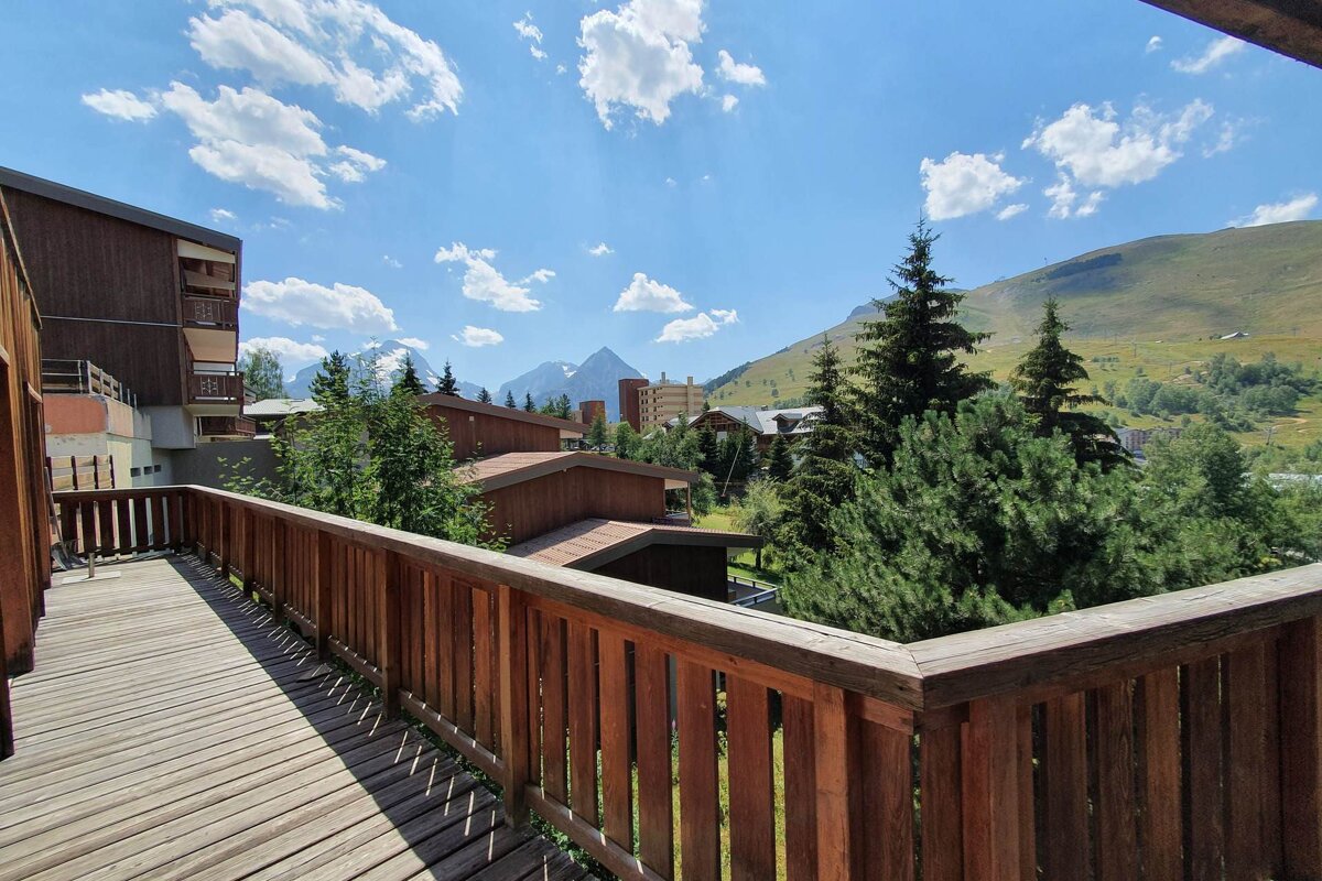 View from a wooden balcony over a mountain village, featuring buildings, pine trees, and distant snow-capped mountains under a sunny, blue sky.