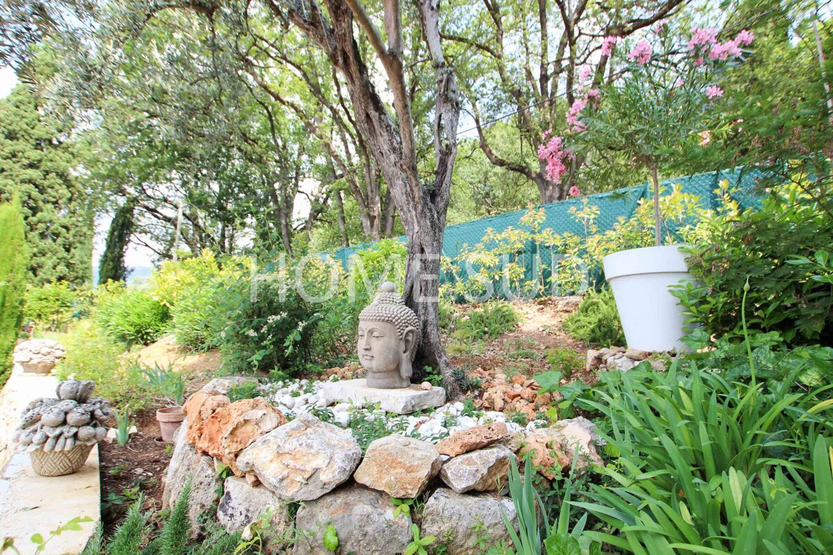 A serene garden with a Buddha head statue nestled among rocks and lush greenery. Trees, pink flowers, and a large white pot complete the natural setting.