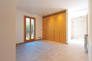 An empty, bright room features a light wood built-in wardrobe, glass French doors, and tiled flooring. A clear view of a shower in an adjacent bathroom is visible.