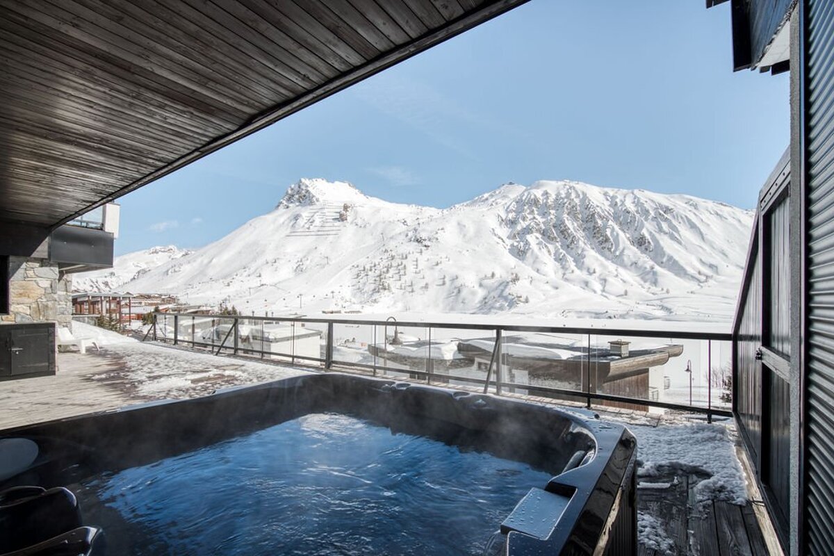 A steaming hot tub on a chalet balcony overlooking majestic snow-covered mountains under a clear blue sky.
