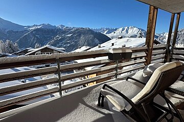 A snow-covered balcony with a lounge chair overlooks a sunny, snow-capped mountain range and alpine chalets under a bright blue sky.