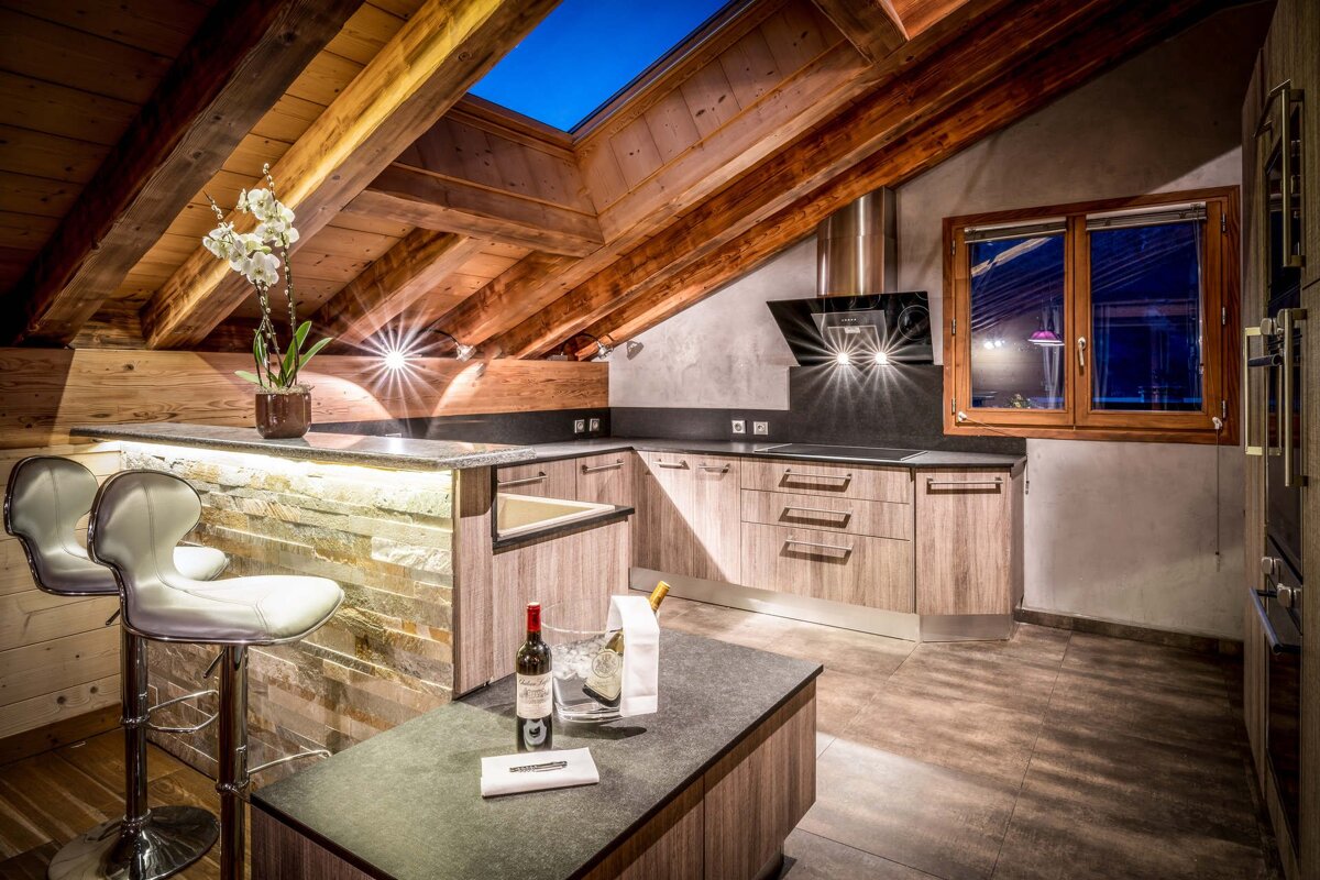 A modern, stylish attic kitchen in a chalet style, featuring exposed wooden beams, a skylight, sleek appliances, and a bar area with wine.
