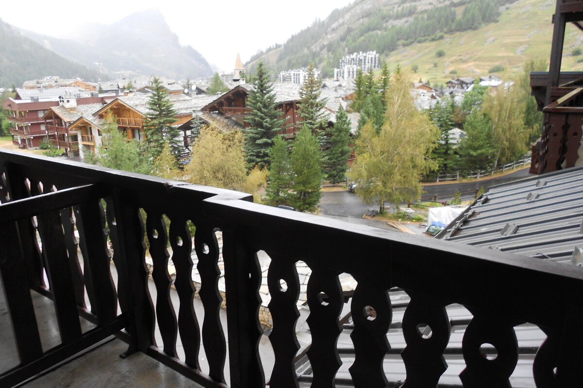 A dark ornate balcony railing frames a misty mountain village with traditional chalets, pine trees, and hazy peaks on an overcast day.