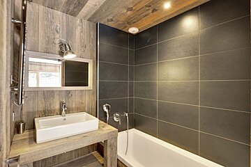 A rustic modern bathroom features wood paneling, a white vessel sink on a wooden vanity, and dark gray tiled walls around a white bathtub.