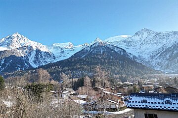 Majestic snow-capped mountains under a clear blue sky overlook a quaint winter village with frosted trees and snowy roofs.