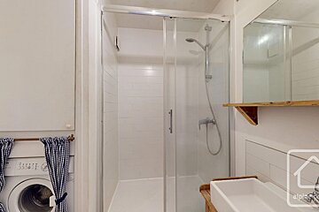 A compact, white bathroom with a glass shower stall, a washing machine covered by a blue checkered curtain, and a sink below a mirror.