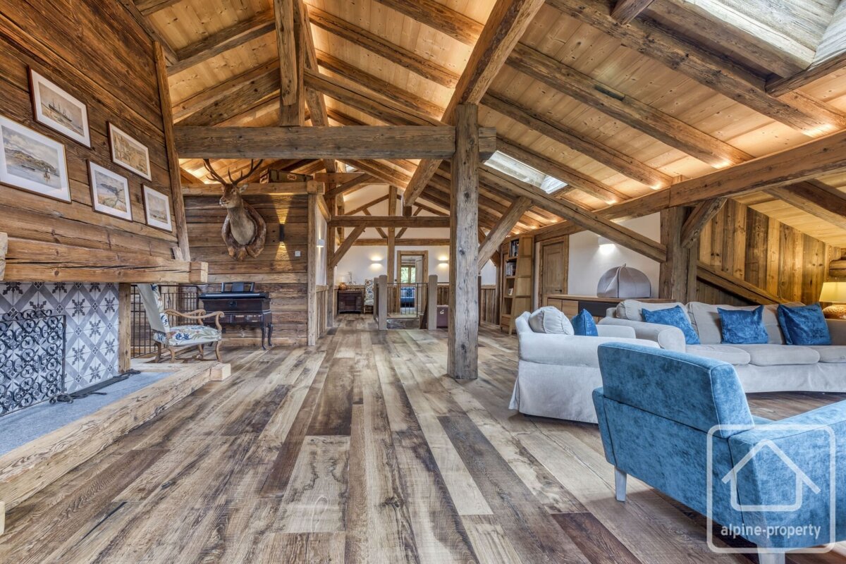 A vast, rustic wooden living room with exposed beams, a tiled fireplace, deer taxidermy, and comfortable sofas with blue cushions.