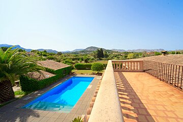A bright blue swimming pool and a tiled rooftop terrace overlook a lush, green landscape with distant mountains under a clear sky.