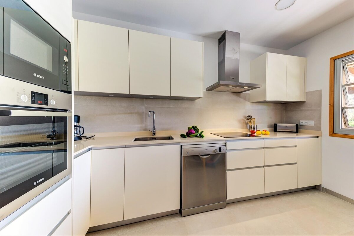 A sleek, modern kitchen with cream cabinets, stainless steel oven, microwave, dishwasher, and range hood. A sink, cooktop, and window complete the bright space.