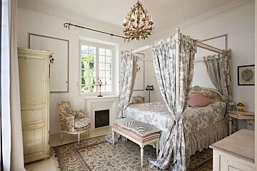 An elegant bedroom with a toile four-poster bed, chandelier, vintage armoire, and window. Features classic decor and a bright ambiance.