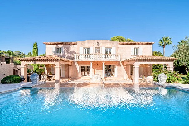 A luxurious pink Mediterranean villa with a sparkling swimming pool in the foreground under a clear blue sky.