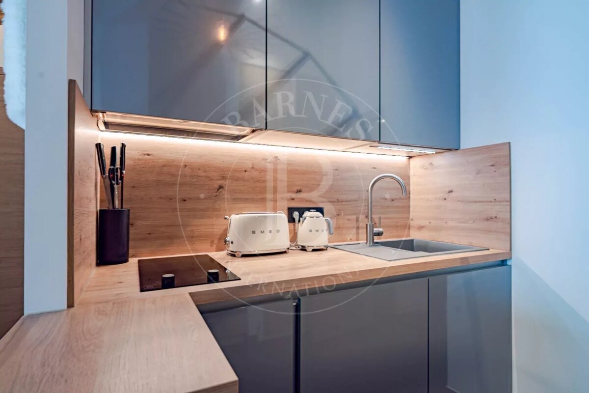 Compact, modern kitchen features glossy gray cabinets, wood accents, under-cabinet lighting, sink, stovetop, and white Smeg appliances.