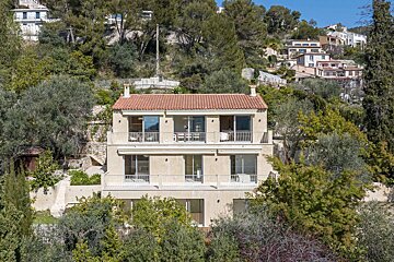 A beige, multi-story house with a red tiled roof and balconies is surrounded by green trees on a sunlit hillside, with other houses in the distance.