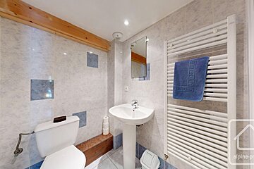 A tiled bathroom featuring a toilet, pedestal sink, mirror, and a white towel warmer with a blue towel. A wooden beam spans the upper wall.