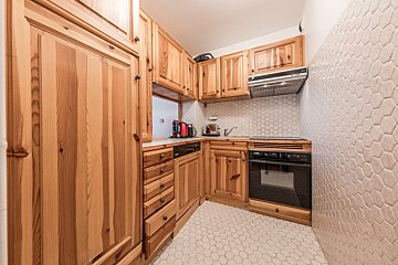 A small kitchen featuring light wood cabinets and hexagonal white tiles on the floor and walls. It includes an oven, stove, sink, and small appliances.