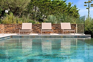 Three modern lounge chairs by a turquoise pool, backed by a stone wall, lush greenery, and a glass fence under a clear sky.