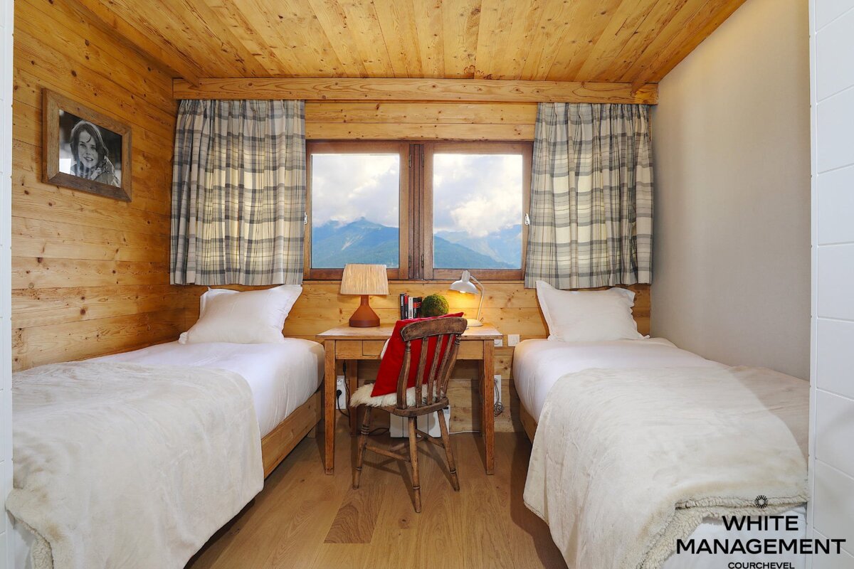 A cozy, rustic wooden bedroom features two single beds, a central desk, and a window framing a stunning mountain view, enhanced by plaid curtains.