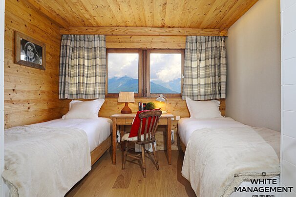 A cozy, rustic wooden bedroom features two single beds, a central desk, and a window framing a stunning mountain view, enhanced by plaid curtains.