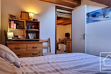 A cozy bedroom with a striped bed, wooden dresser/bookshelf, and a lamp. An open doorway reveals another room with a TV. Wooden beams accent the ceiling.