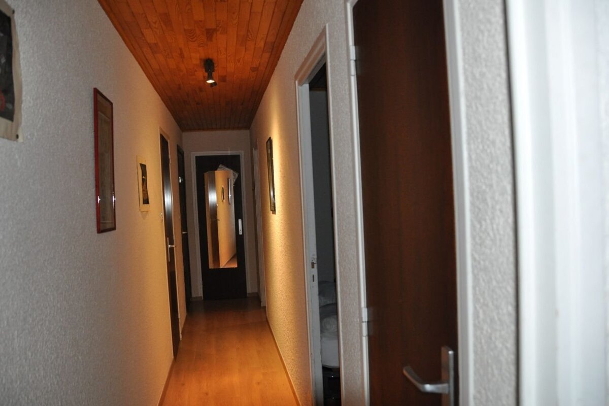 Narrow hallway with white walls, a wooden plank ceiling, doors on both sides, and a door with a mirror at the far end.