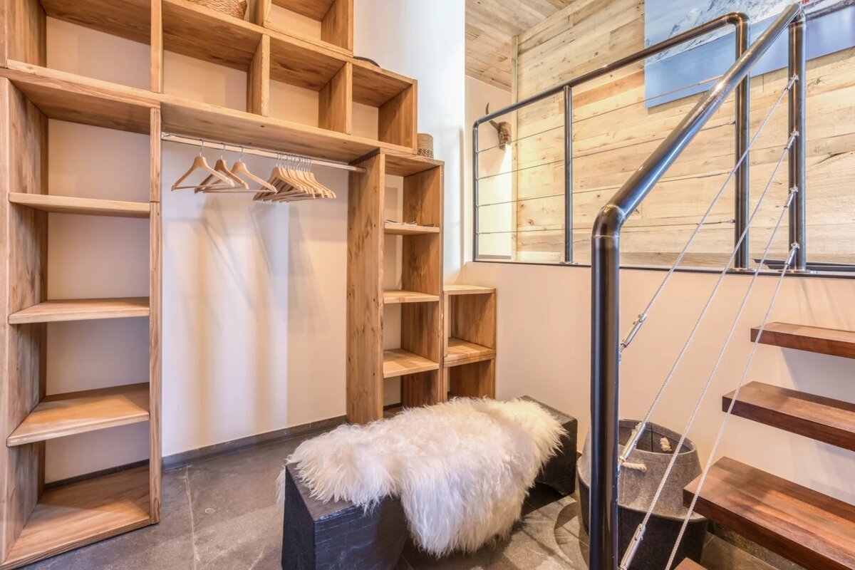 A rustic entryway featuring an open wooden closet, a bench with a white fur throw, and floating stairs.