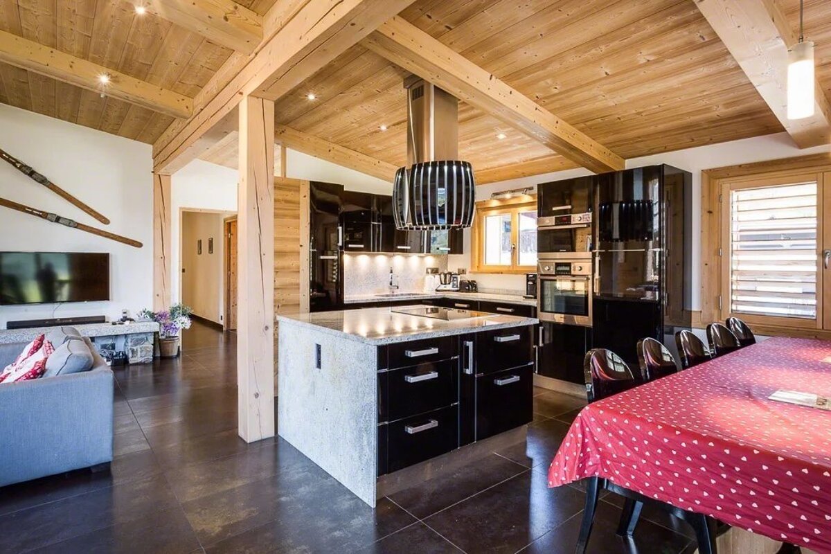A modern chalet kitchen and living area with exposed wooden beams, sleek black cabinets, and a dining table with a red tablecloth.