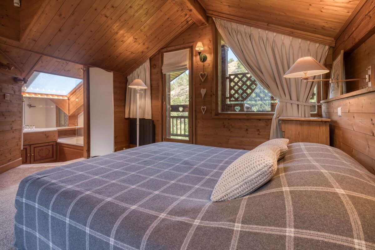 Cozy, rustic wood-paneled bedroom featuring a plaid bed, mountain view window, and an en-suite bathroom with a skylight visible through an archway.