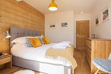 Cozy bedroom with a wood-paneled accent wall, a grey upholstered bed with yellow pillows, and warm wooden furniture.