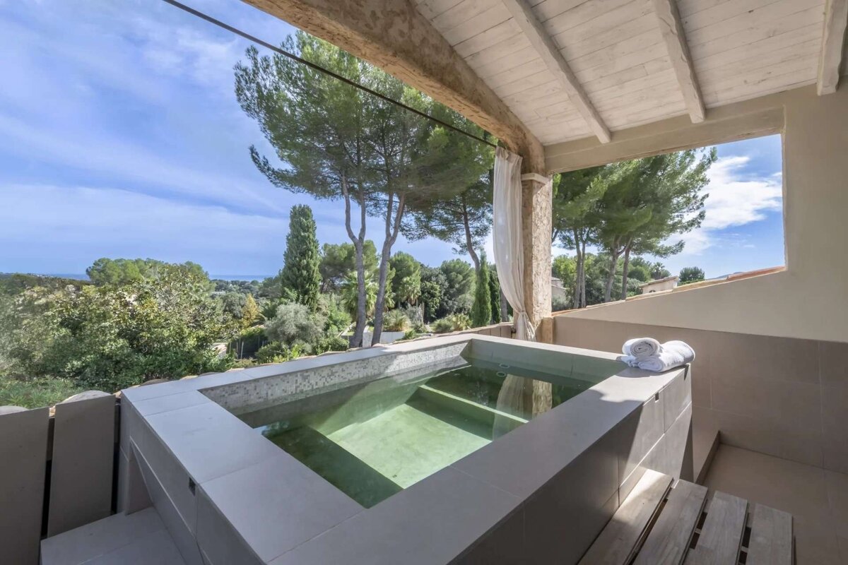 A covered balcony with a built-in hot tub overlooks a lush, green landscape with trees and a distant view of the sea under a blue sky.