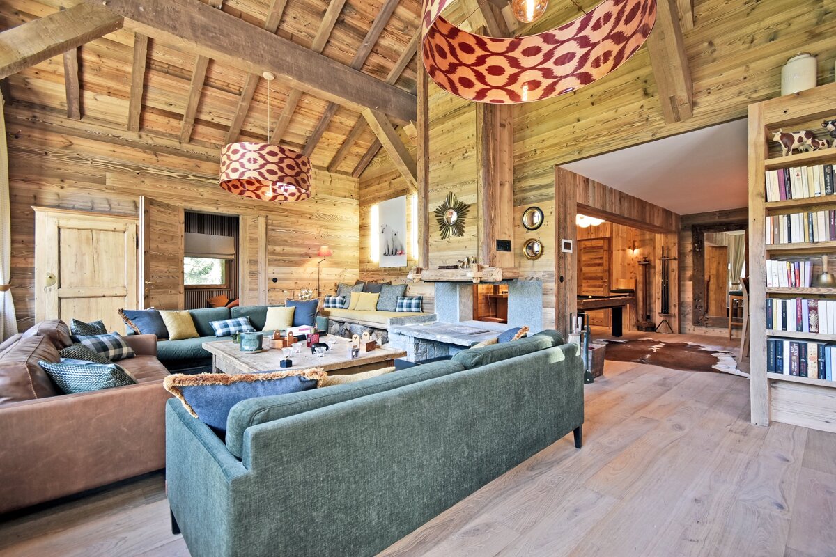 A warm, wood-paneled chalet living room featuring several inviting sofas, a fireplace, and a unique patterned chandelier.