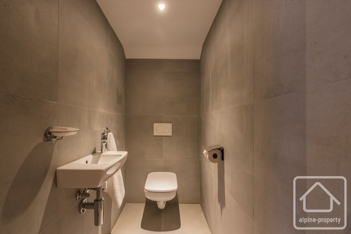 A modern, minimalist toilet room with gray tiled walls, a wall-mounted white toilet, and a small sink with a hand towel.