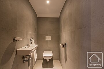 A modern, minimalist toilet room with gray tiled walls, a wall-mounted white toilet, and a small sink with a hand towel.