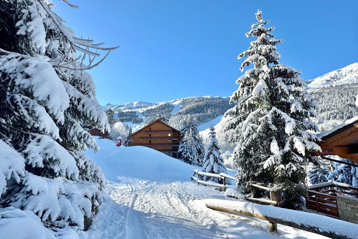 A vibrant winter wonderland, showcasing snow-laden pine trees, wooden chalets, and majestic mountains under a clear blue sky. Ski slopes are visible in the background.