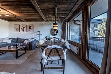 A rustic chalet interior with a wooden dining table, a cozy living area with a grey sectional, and large windows.