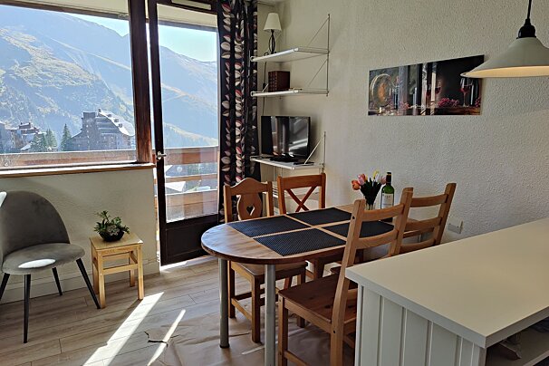 Bright dining area with a wooden table, chairs, TV, and a balcony offering a scenic mountain view with distant buildings. Flowers and wine on table.