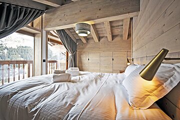 Cozy wooden chalet bedroom featuring a large white bed, warm reading lights, and balcony views of snowy mountains.