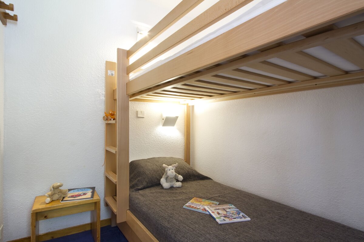 A cozy room featuring wooden bunk beds. The lower bunk has a gray blanket, a plush toy, and books, lit by a wall lamp. A small table holds another toy.