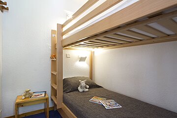 A cozy room featuring wooden bunk beds. The lower bunk has a gray blanket, a plush toy, and books, lit by a wall lamp. A small table holds another toy.