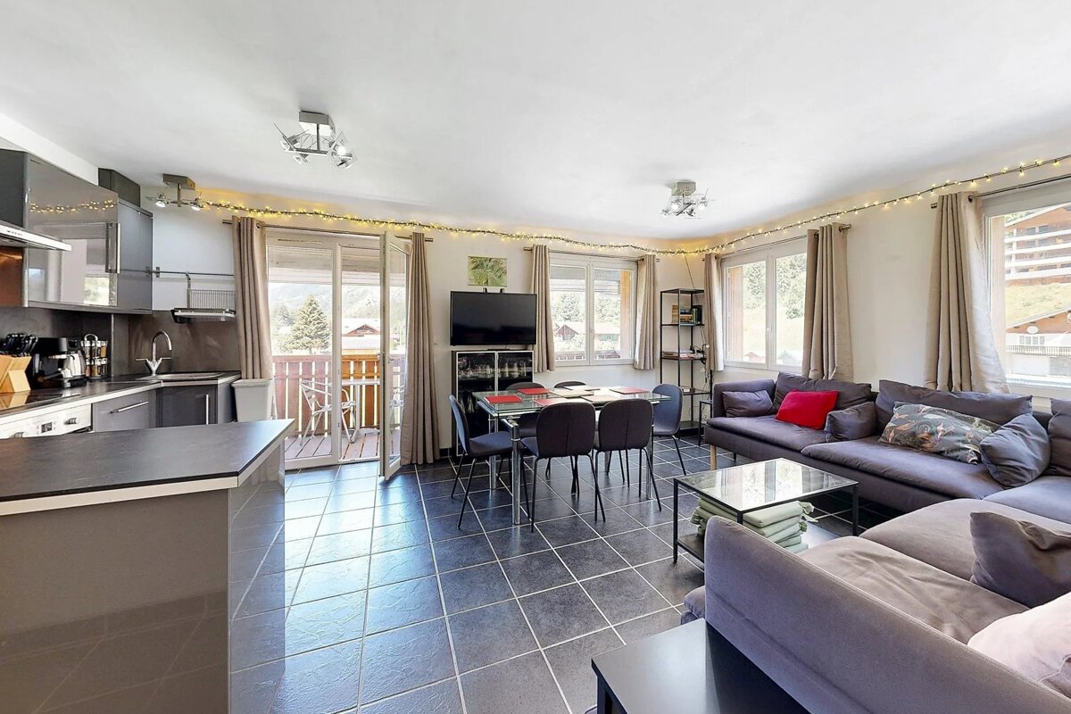 Bright open-plan living space featuring a kitchen, dining table, gray sectional sofa, tiled floor, and balcony doors.