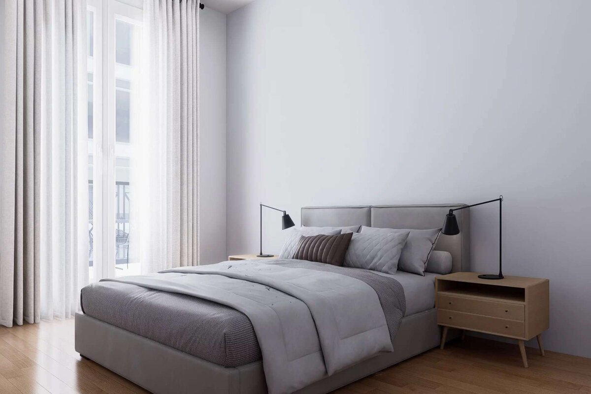 A modern, minimalist bedroom features a gray bed with light bedding, wooden nightstands and lamps, and a large window with flowing white curtains.