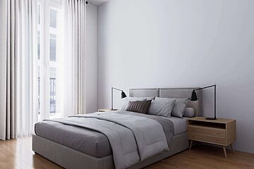 A modern, minimalist bedroom features a gray bed with light bedding, wooden nightstands and lamps, and a large window with flowing white curtains.