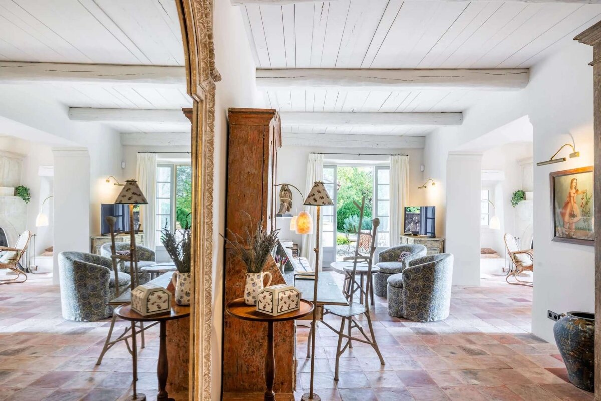 Bright, rustic living room with terracotta floors, white beamed ceilings, antique wooden furniture, and lamps. Large windows overlook a serene garden.