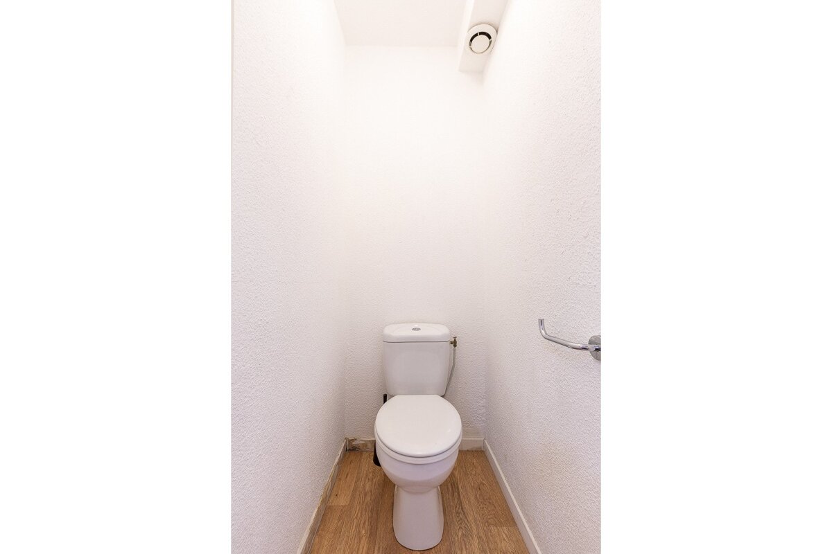 A white toilet stands in a narrow room with textured white walls, a light wooden floor, a ceiling vent, and a chrome toilet paper holder.