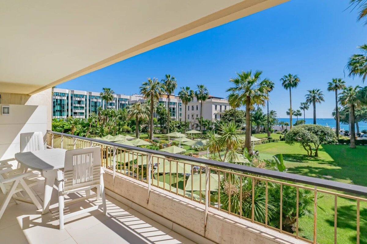 Sunny resort balcony overlooking a lush green garden with palm trees, umbrellas, and a bright blue sky.